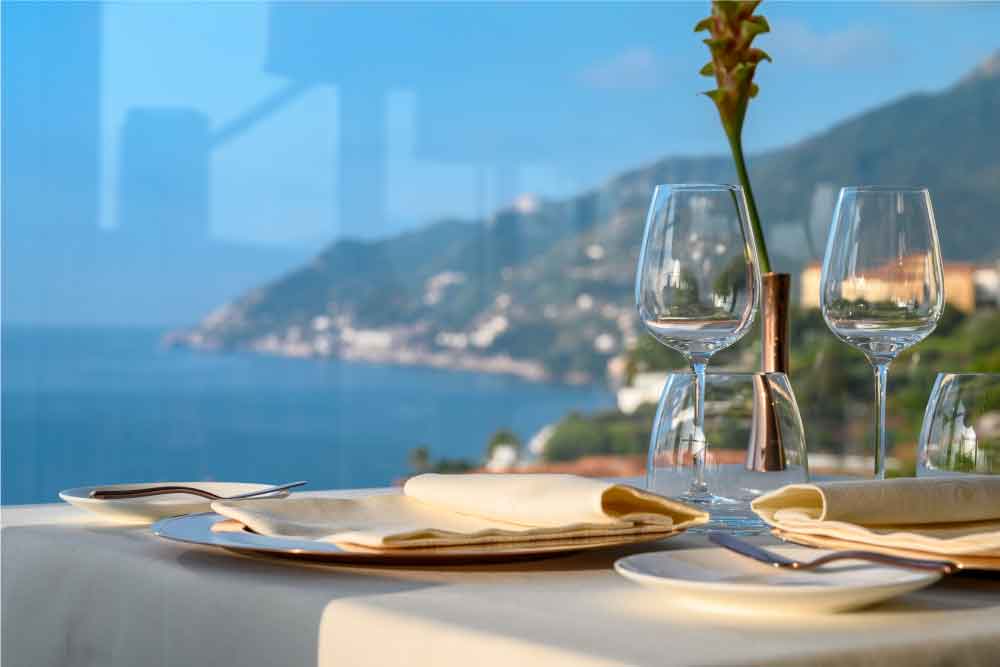 Mesa posta com taças de vidro junto a uma janela, vista panorâmica do mar e da costa ao fundo.