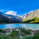 Uma paisagem de tirar o fôlego com um lago de águas azul-turquesa cercado por montanhas altas e rochosas, com o céu azul e algumas nuvens brancas ao fundo.