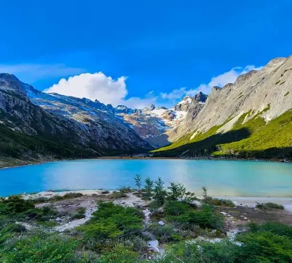 Uma paisagem de tirar o fôlego com um lago de águas azul-turquesa cercado por montanhas altas e rochosas, com o céu azul e algumas nuvens brancas ao fundo.