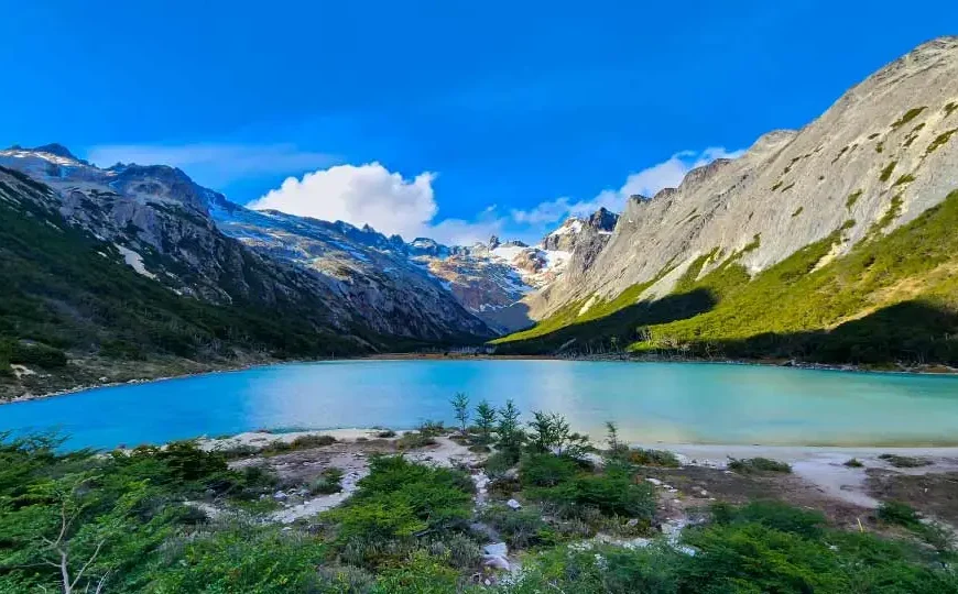 Uma paisagem de tirar o fôlego com um lago de águas azul-turquesa cercado por montanhas altas e rochosas, com o céu azul e algumas nuvens brancas ao fundo.