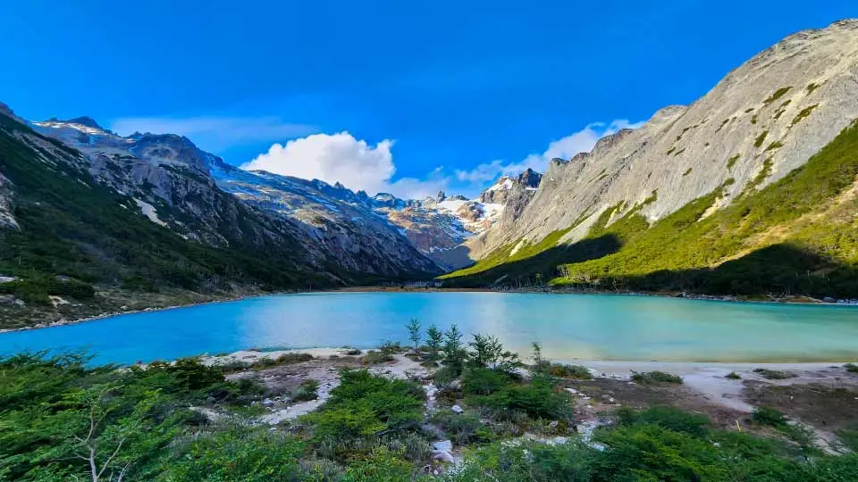 Uma paisagem de tirar o fôlego com um lago de águas azul-turquesa cercado por montanhas altas e rochosas, com o céu azul e algumas nuvens brancas ao fundo.