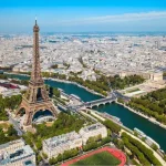 Paisagem aérea do rio Sena com a Torre Eiffel ao centro, cercada por áreas verdes, pontes e a cidade de Paris ao fundo, sob céu azul claro.