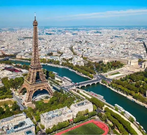 Paisagem aérea do rio Sena com a Torre Eiffel ao centro, cercada por áreas verdes, pontes e a cidade de Paris ao fundo, sob céu azul claro.