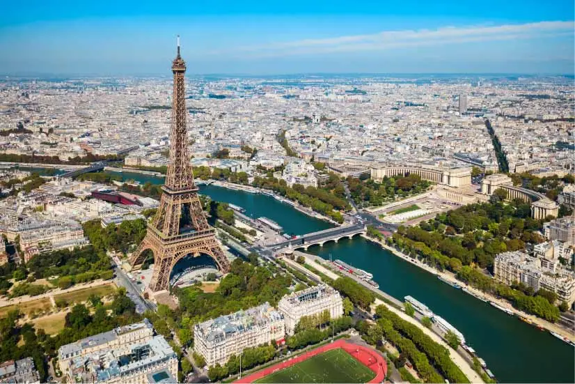 Paisagem aérea do rio Sena com a Torre Eiffel ao centro, cercada por áreas verdes, pontes e a cidade de Paris ao fundo, sob céu azul claro.