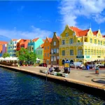 Casario colorido ao longo de um canal, com edifícios em tons vibrantes, cafés ao ar livre com guarda-sóis brancos e um céu azul com algumas nuvens.