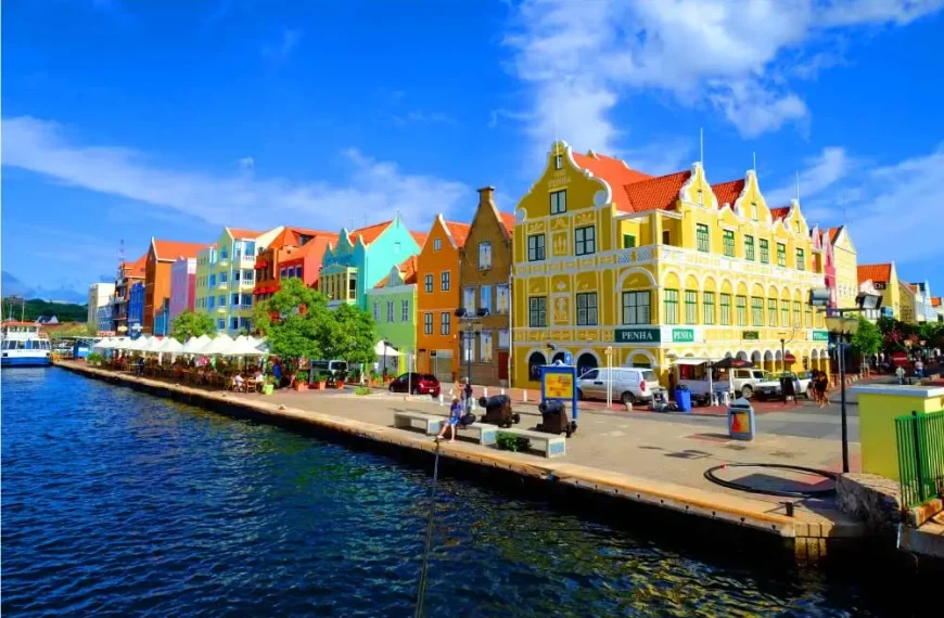 Casario colorido ao longo de um canal, com edifícios em tons vibrantes, cafés ao ar livre com guarda-sóis brancos e um céu azul com algumas nuvens.