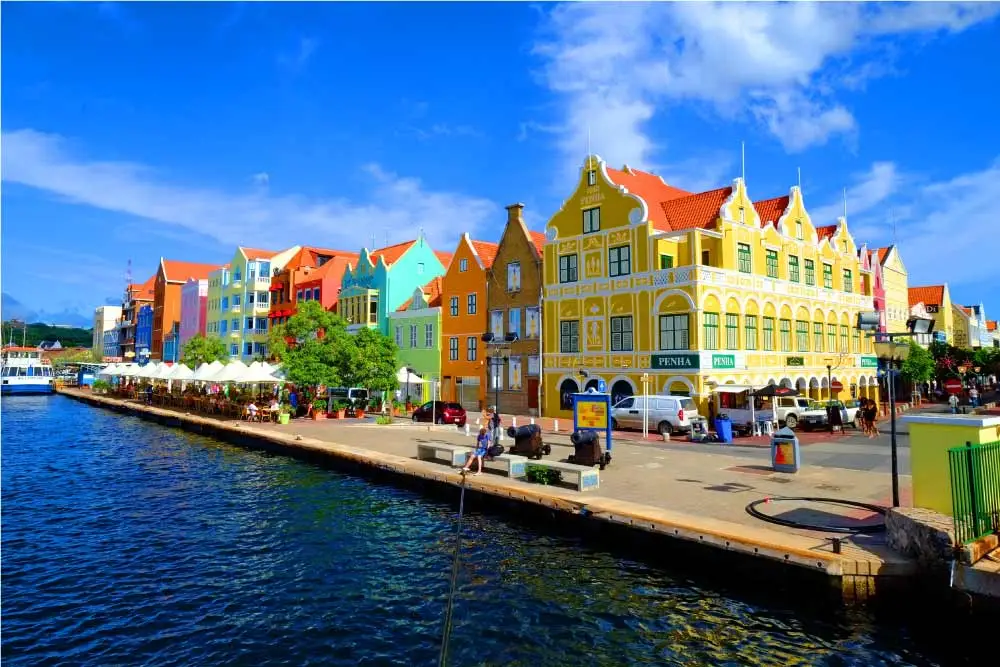 Casario colorido ao longo de um canal, com edifícios em tons vibrantes, cafés ao ar livre com guarda-sóis brancos e um céu azul com algumas nuvens.