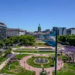 Bandeira argentina esvoaçando com o prédio do Congresso Nacional e um parque paisagístico ao fundo em Buenos Aires, Argentina. A cena é vista de cima e mostra edifícios circundantes.