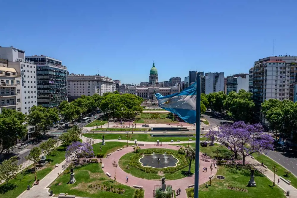Bandeira argentina esvoaçando com o prédio do Congresso Nacional e um parque paisagístico ao fundo em Buenos Aires, Argentina. A cena é vista de cima e mostra edifícios circundantes.