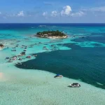 Vista aérea de ilha e bancos de areia em águas turquesa com vários barcos ancorados.