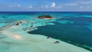 Vista aérea de ilha e bancos de areia em águas turquesa com vários barcos ancorados.