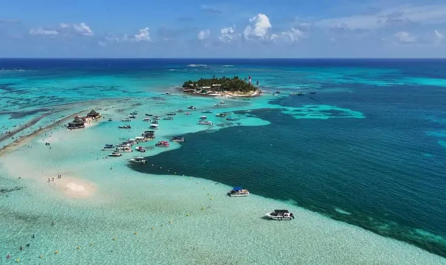 Vista aérea de ilha e bancos de areia em águas turquesa com vários barcos ancorados.