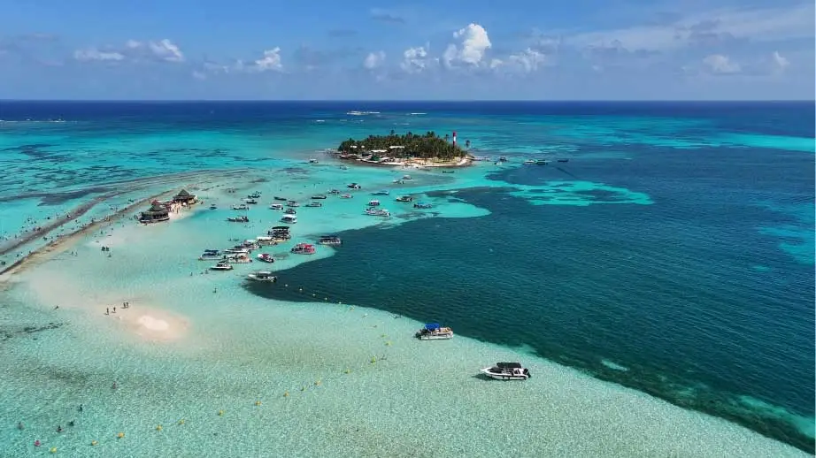 Vista aérea de ilha e bancos de areia em águas turquesa com vários barcos ancorados.