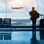 Pessoa de costas segurando uma caneca e a alça de uma mala de rodinhas, parada diante de uma grande parede de vidro do terminal; ao fundo vê-se um avião em voo sobre a pista e hangares sob um céu nublado, com cadeiras vazias ao lado e reflexos no piso brilhante.