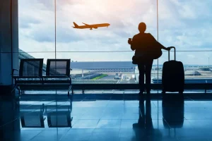 Pessoa de costas segurando uma caneca e a alça de uma mala de rodinhas, parada diante de uma grande parede de vidro do terminal; ao fundo vê-se um avião em voo sobre a pista e hangares sob um céu nublado, com cadeiras vazias ao lado e reflexos no piso brilhante.