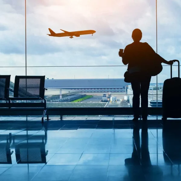Pessoa de costas segurando uma caneca e a alça de uma mala de rodinhas, parada diante de uma grande parede de vidro do terminal; ao fundo vê-se um avião em voo sobre a pista e hangares sob um céu nublado, com cadeiras vazias ao lado e reflexos no piso brilhante.