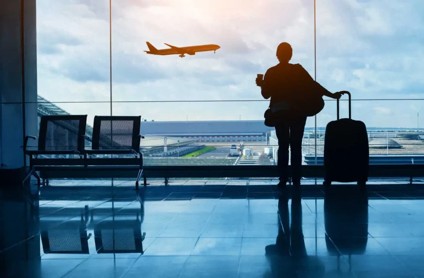 Pessoa de costas segurando uma caneca e a alça de uma mala de rodinhas, parada diante de uma grande parede de vidro do terminal; ao fundo vê-se um avião em voo sobre a pista e hangares sob um céu nublado, com cadeiras vazias ao lado e reflexos no piso brilhante.