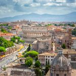 Vista panorâmica do centro de Roma, com o Coliseu ao fundo e ruas movimentadas com árvores verdes e edifícios históricos sob céu parcialmente nublado