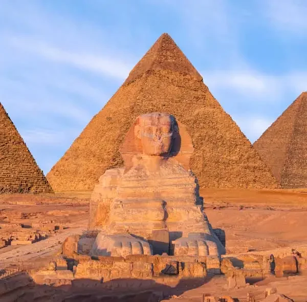 Imagem de um sítio arqueológico no Egito, mostrando as Pirâmides de Gizé ao fundo e a Esfinge à frente, sob um céu parcialmente nublado.