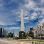 Imagem da praça do obelisco em Buenos Aires, com arbustos bem cuidados formando figuras geométricas, e prédios históricos ao redor.