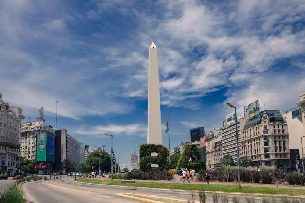 Imagem da praça do obelisco em Buenos Aires, com arbustos bem cuidados formando figuras geométricas, e prédios históricos ao redor.