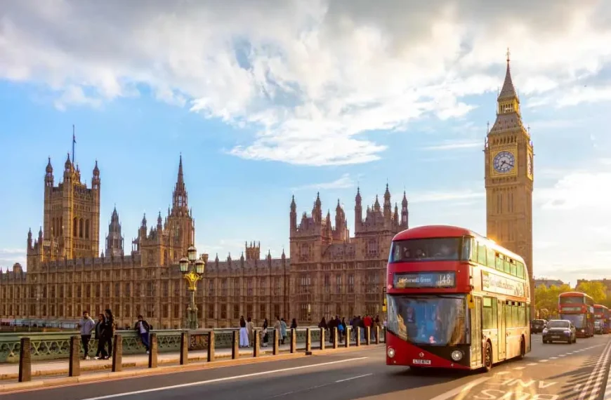 Imagem de um ônibus de dois andares vermelho e verde no lado de uma ponte na cidade de Londres, com o Big Ben e o Palácio de Westminster ao fundo.