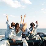 Quatro amigos, de costas, comemorando em um carro conversível próximo ao mar, com os braços levantados.