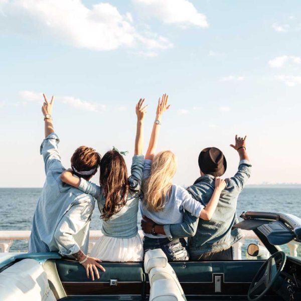 Quatro amigos, de costas, comemorando em um carro conversível próximo ao mar, com os braços levantados.