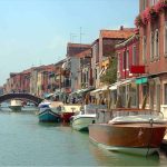 Uma vista panorâmica de um canal em Veneza, Itália, ladeado por edifícios coloridos e barcos atracados. No fundo, uma ponte arqueada atravessa o canal e uma torre de igreja pode ser vista. O céu está azul claro.