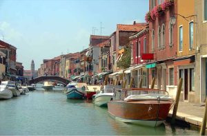 Uma vista panorâmica de um canal em Veneza, Itália, ladeado por edifícios coloridos e barcos atracados. No fundo, uma ponte arqueada atravessa o canal e uma torre de igreja pode ser vista. O céu está azul claro.