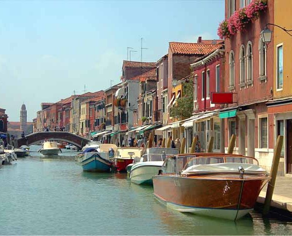 Uma vista panorâmica de um canal em Veneza, Itália, ladeado por edifícios coloridos e barcos atracados. No fundo, uma ponte arqueada atravessa o canal e uma torre de igreja pode ser vista. O céu está azul claro.