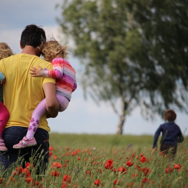 Viagem em família, pai segurando duas filhas e filho a frente brincando em um campo