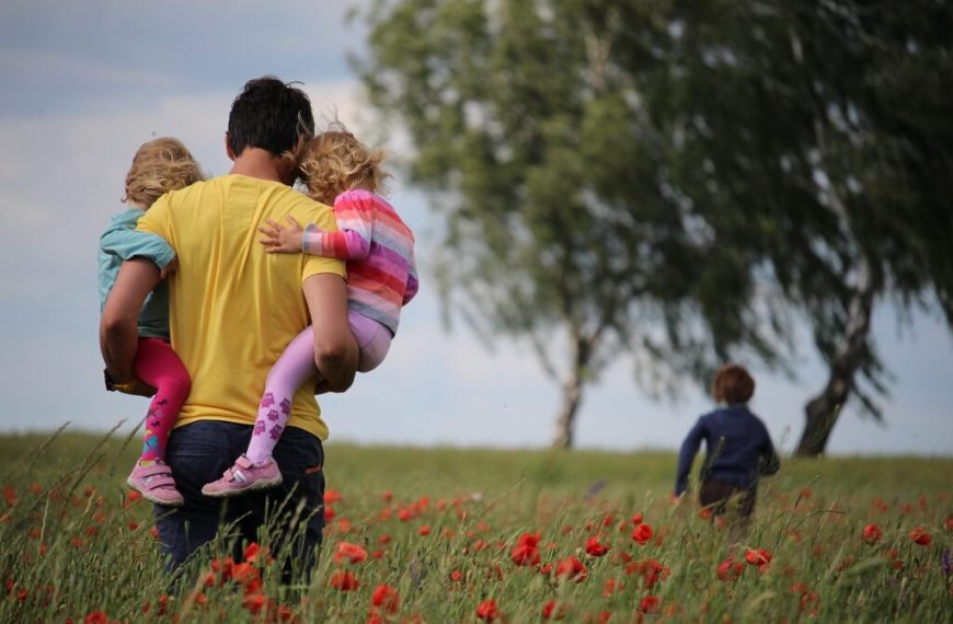 Viagem em família, pai segurando duas filhas e filho a frente brincando em um campo