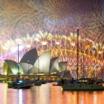 Uma exibição espetacular de fogos de artifício ilumina o céu noturno acima da Ópera de Sydney e da Ponte da Baía de Sydney, com vários barcos ancorados na água em primeiro plano.