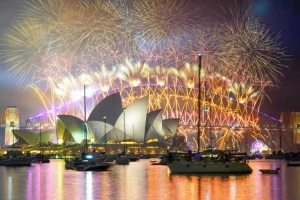 Uma exibição espetacular de fogos de artifício ilumina o céu noturno acima da Ópera de Sydney e da Ponte da Baía de Sydney, com vários barcos ancorados na água em primeiro plano.