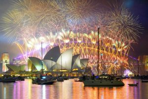Uma exibição espetacular de fogos de artifício ilumina o céu noturno acima da Ópera de Sydney e da Ponte da Baía de Sydney, com vários barcos ancorados na água em primeiro plano.