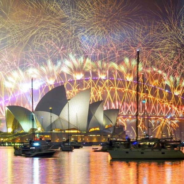 Uma exibição espetacular de fogos de artifício ilumina o céu noturno acima da Ópera de Sydney e da Ponte da Baía de Sydney, com vários barcos ancorados na água em primeiro plano.
