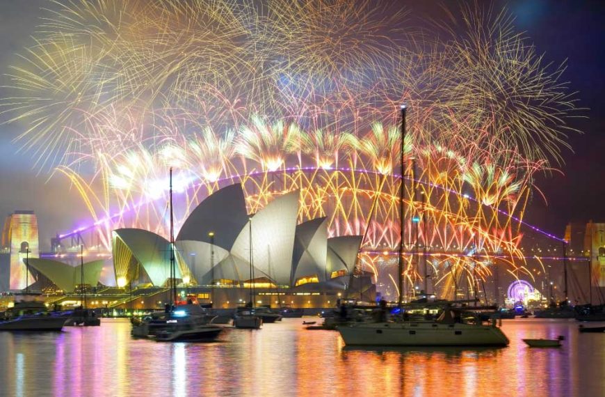 Uma exibição espetacular de fogos de artifício ilumina o céu noturno acima da Ópera de Sydney e da Ponte da Baía de Sydney, com vários barcos ancorados na água em primeiro plano.
