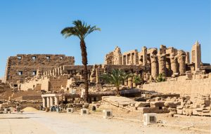 Vista de um templo antigo e suas ruínas, com paredes de pedra e colunas altas sob um céu azul claro. Uma palmeira alta se eleva no centro, e algumas palmeiras menores são visíveis entre as ruínas. O chão é de terra batida com algumas lajes de pedra, e há algumas pequenas estruturas retangulares brancas e caixas no primeiro plano.