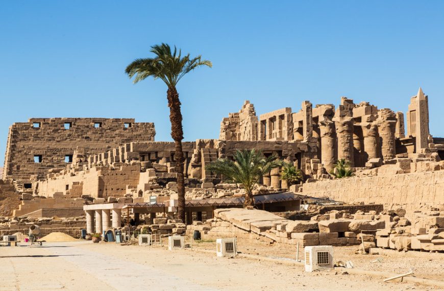 Vista de um templo antigo e suas ruínas, com paredes de pedra e colunas altas sob um céu azul claro. Uma palmeira alta se eleva no centro, e algumas palmeiras menores são visíveis entre as ruínas. O chão é de terra batida com algumas lajes de pedra, e há algumas pequenas estruturas retangulares brancas e caixas no primeiro plano.