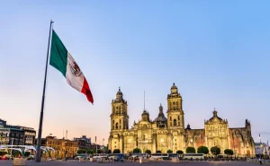 A bandeira do México tremulando em primeiro plano, com a Catedral Metropolitana da Cidade do México iluminada ao entardecer no fundo, em uma praça movimentada.