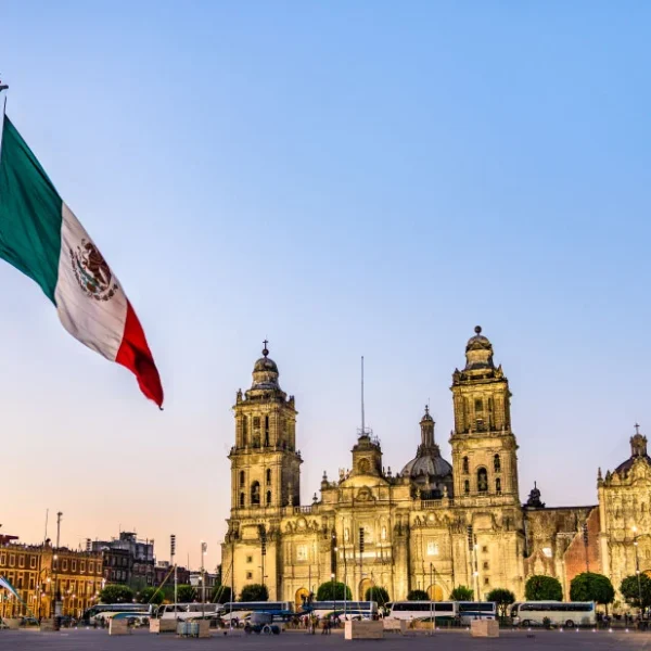 A bandeira do México tremulando em primeiro plano, com a Catedral Metropolitana da Cidade do México iluminada ao entardecer no fundo, em uma praça movimentada.
