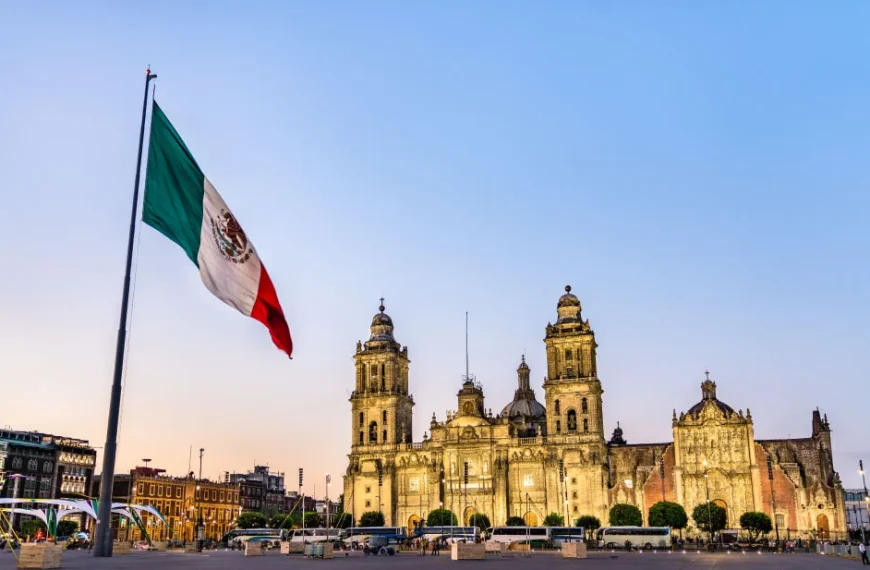 A bandeira do México tremulando em primeiro plano, com a Catedral Metropolitana da Cidade do México iluminada ao entardecer no fundo, em uma praça movimentada.
