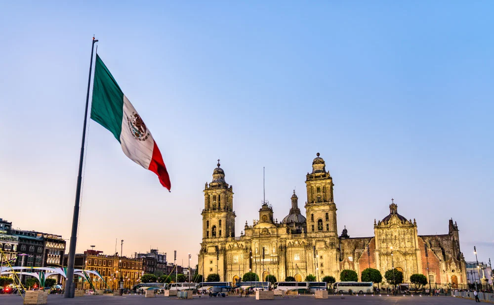 A bandeira do México tremulando em primeiro plano, com a Catedral Metropolitana da Cidade do México iluminada ao entardecer no fundo, em uma praça movimentada.