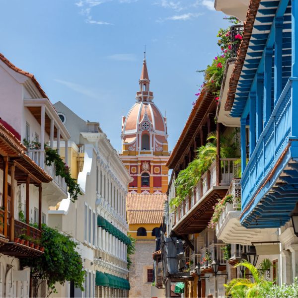 Uma rua estreita e ensolarada em Cartagena, Colômbia, ladeada por edifícios coloniais brancos e coloridos com varandas de madeira. No centro, ao fundo, ergue-se a cúpula e a torre de uma igreja em tons de terracota e branco. As varandas estão decoradas com plantas e flores, e o céu azul claro é visível acima.
