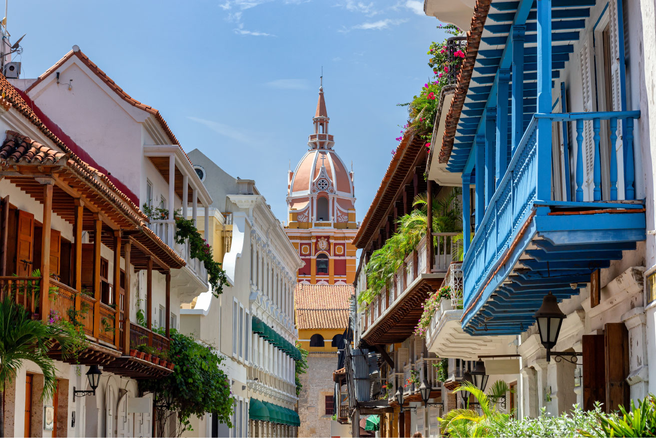 Uma rua estreita e ensolarada em Cartagena, Colômbia, ladeada por edifícios coloniais brancos e coloridos com varandas de madeira. No centro, ao fundo, ergue-se a cúpula e a torre de uma igreja em tons de terracota e branco. As varandas estão decoradas com plantas e flores, e o céu azul claro é visível acima.