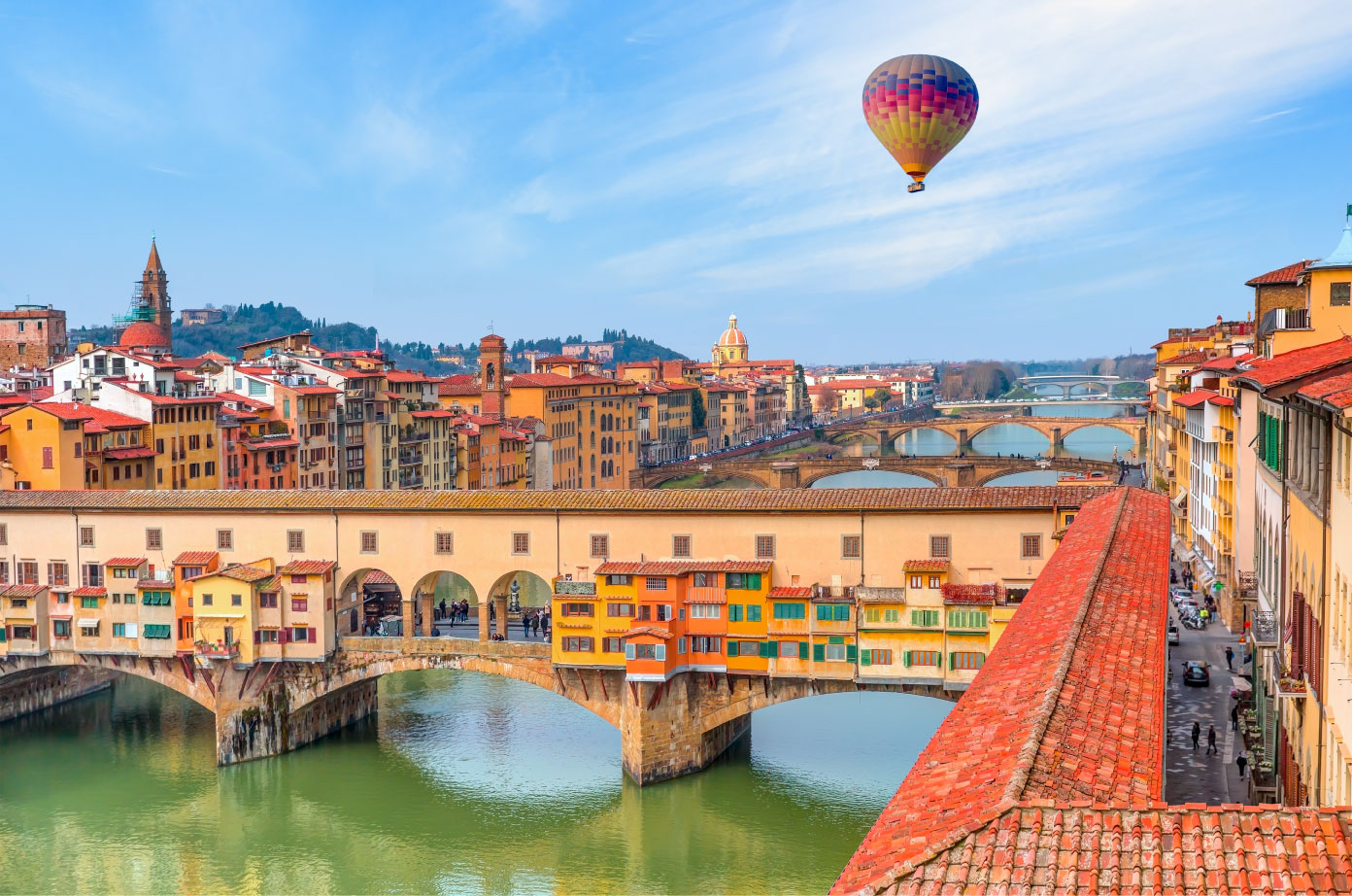 Uma vista panorâmica da Ponte Vecchio e dos edifícios coloridos ao longo do Rio Arno em Florença, Itália, com um balão de ar quente subindo no céu azul.
