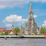 Vista do Templo Wat Arun às margens do Rio Chao Phraya, em Bangkok, com sua torre central ornamentada destacando-se contra um céu azul com nuvens, rodeado por edifícios coloridos e bandeiras tailandesas.