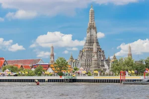 Vista do Templo Wat Arun às margens do Rio Chao Phraya, em Bangkok, com sua torre central ornamentada destacando-se contra um céu azul com nuvens, rodeado por edifícios coloridos e bandeiras tailandesas.