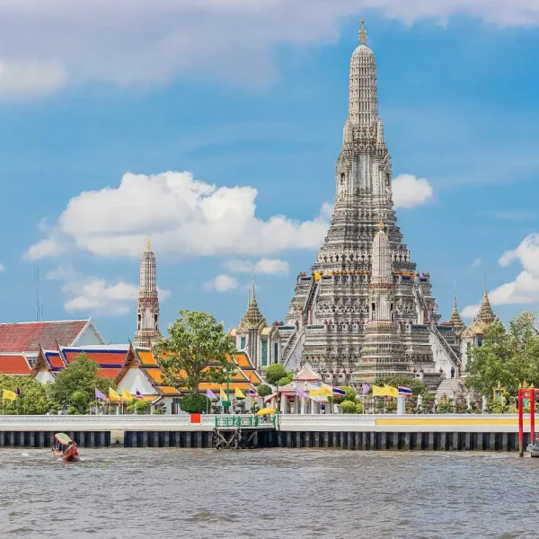 Vista do Templo Wat Arun às margens do Rio Chao Phraya, em Bangkok, com sua torre central ornamentada destacando-se contra um céu azul com nuvens, rodeado por edifícios coloridos e bandeiras tailandesas.